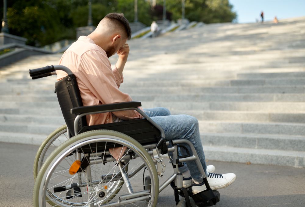 Chico triste en silla de ruedas al pie de unas escaleras. Especialidad tratada para adaptación y convivencia con enfermedades físicas.
