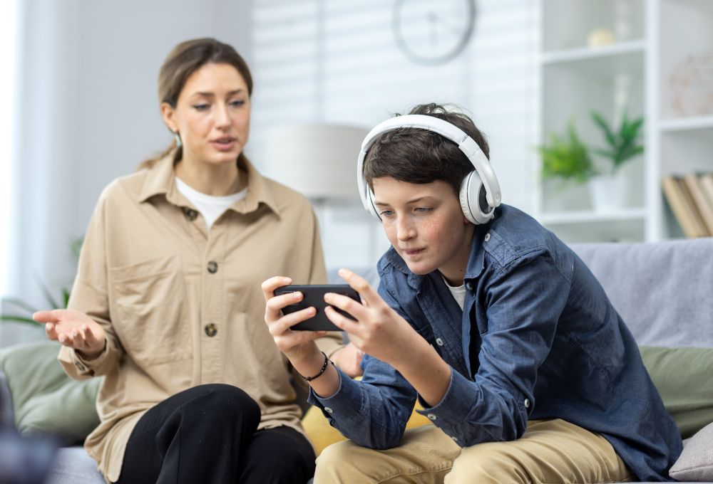 Conflicto familiar porque la madre riñe con su hijo, adolescente con auriculares, por estar siempre jugando en el móvil. Concepto de adicciones al móvil y otras tecnologías.